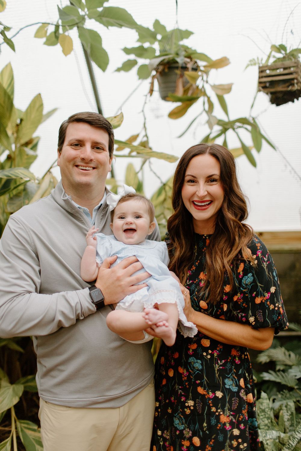 Family poses together in greenhouse setting wearing coordinated outfits with floral dress and gray shirt.