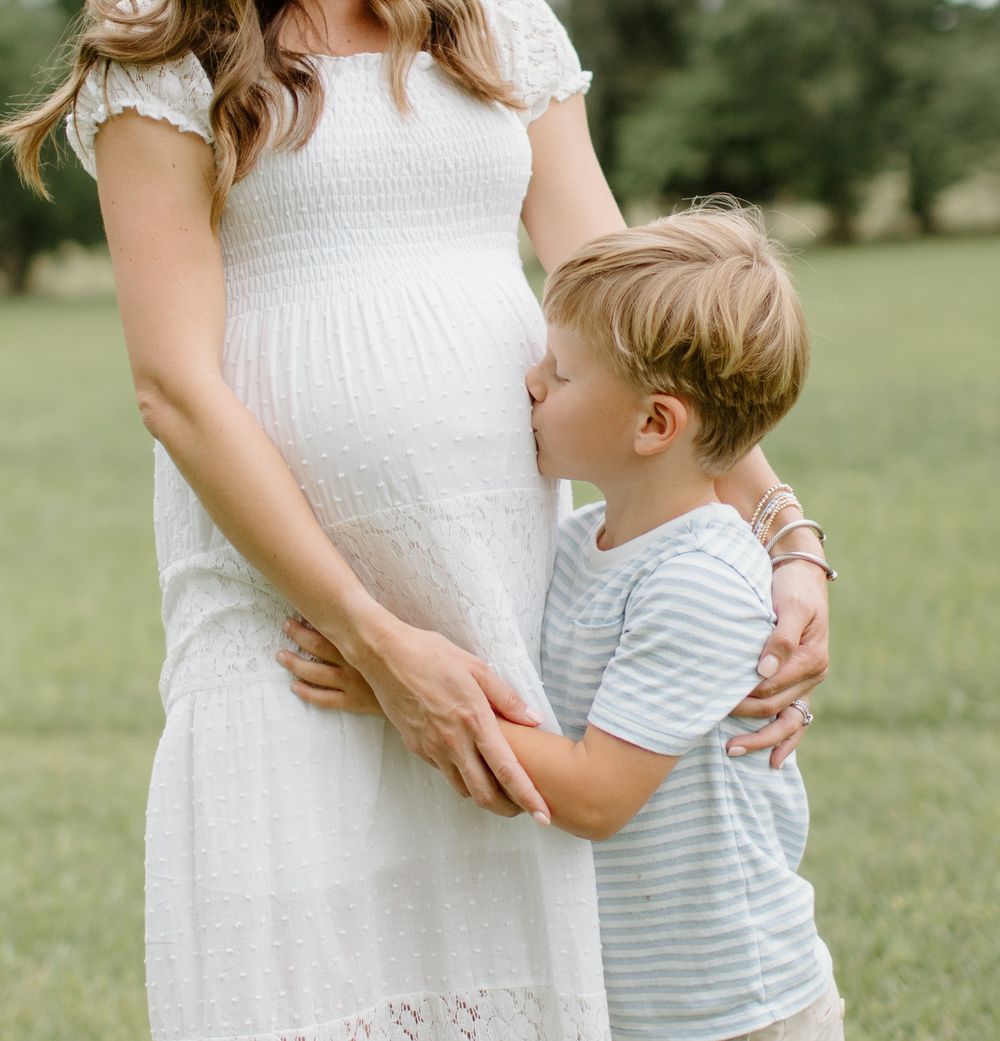 Mother in white dress embraces young child in striped shirt outdoors.