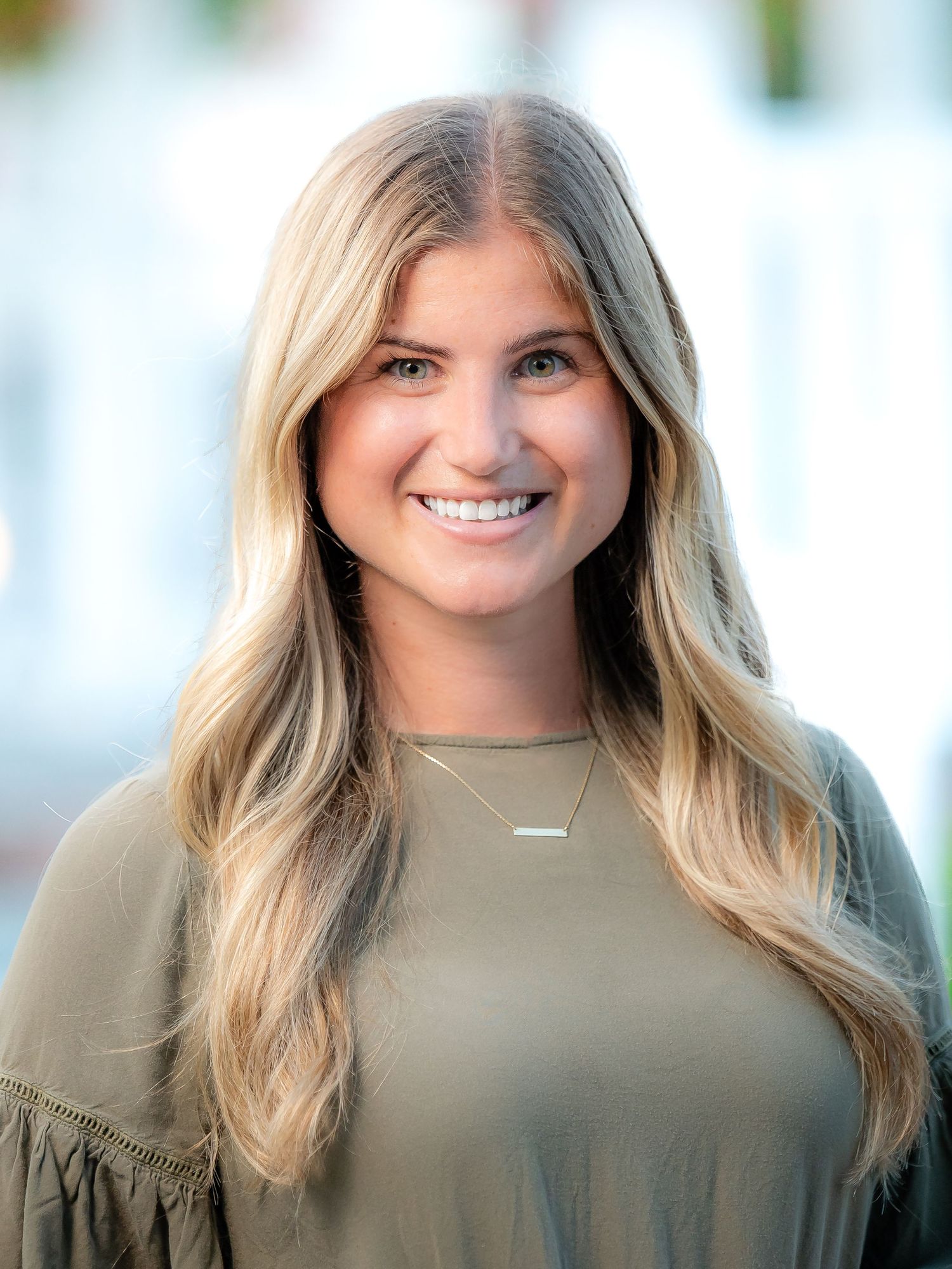 Professional portrait of someone wearing an olive green top with flowing blonde hair and a delicate necklace in natural light.