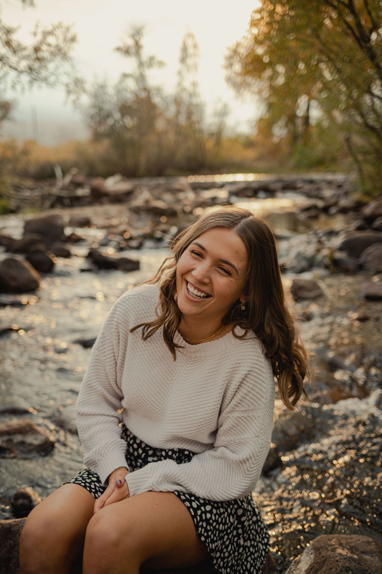 aurora senior photo of someone sitting on rocks by a stream at sunset wearing a cozy sweater