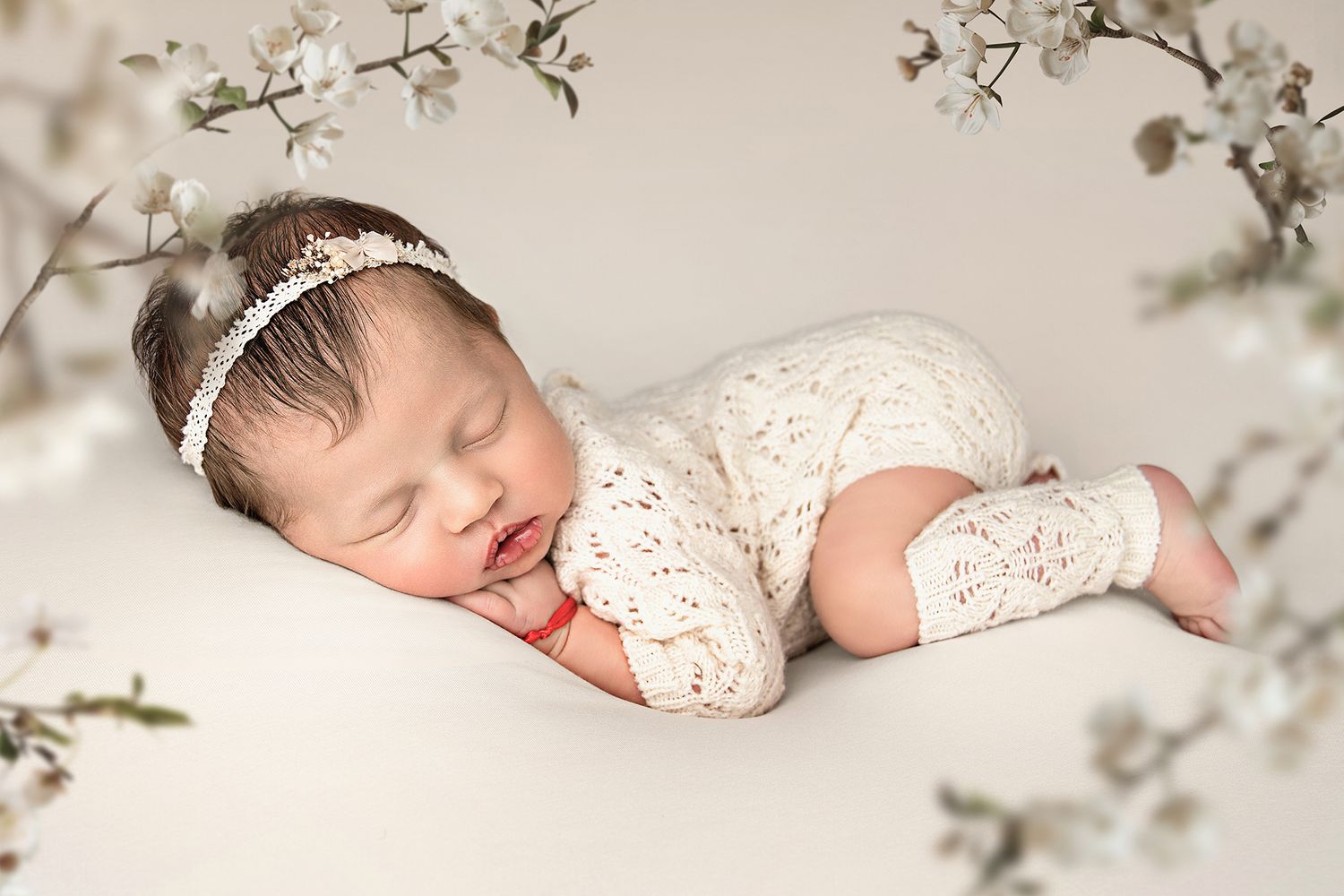 A sleeping baby wearing white lace lies peacefully among delicate cherry blossoms and soft floral arrangements.
