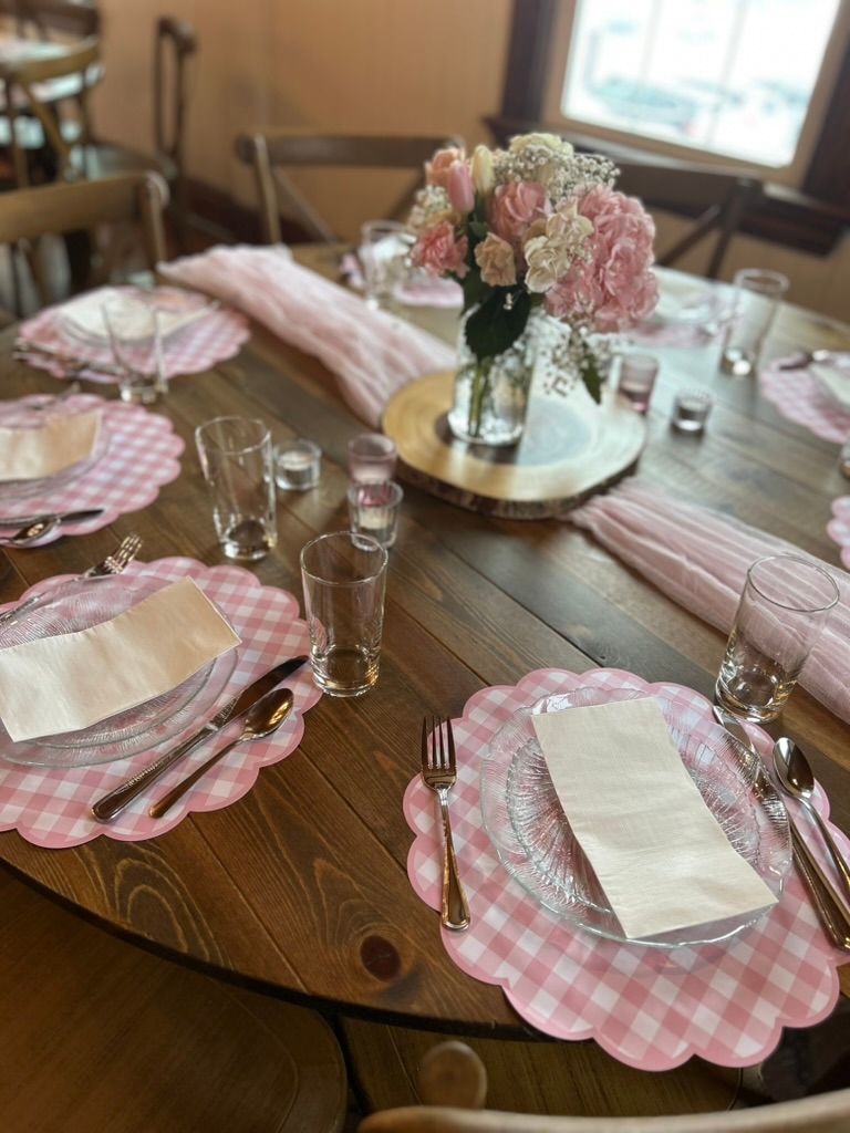 Pink gingham placemat table setting with pink roses, hydrangeas, clear glassware, and rose gold cutlery on a round wood table.