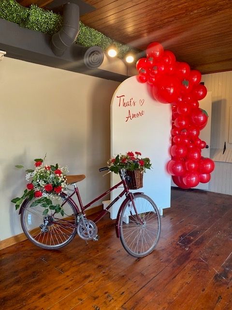 Italian-themed party backdrop with red balloon arch, decorative bicycle with roses, and That's Amore signage on white arch.