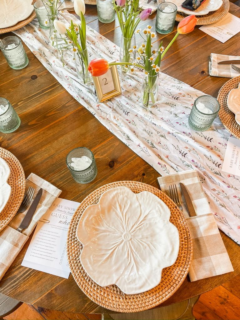 Spring bridal shower table with floral runner, wildflower bud vases, rattan chargers, and scalloped white plates overhead view.
