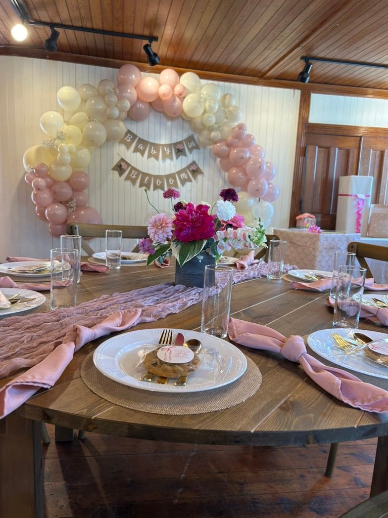 Pink baby shower table with balloon garland backdrop, floral centerpiece, pink velvet runner, and Happy Birthday banner.