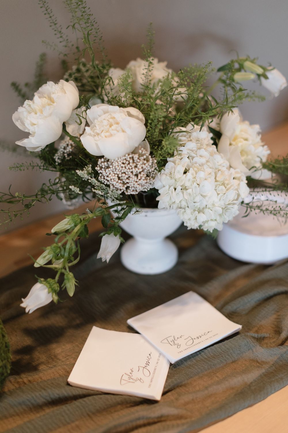 White floral arrangement of peonies and hydrangeas in a milk glass vase beside handwritten calligraphy place cards on a table.