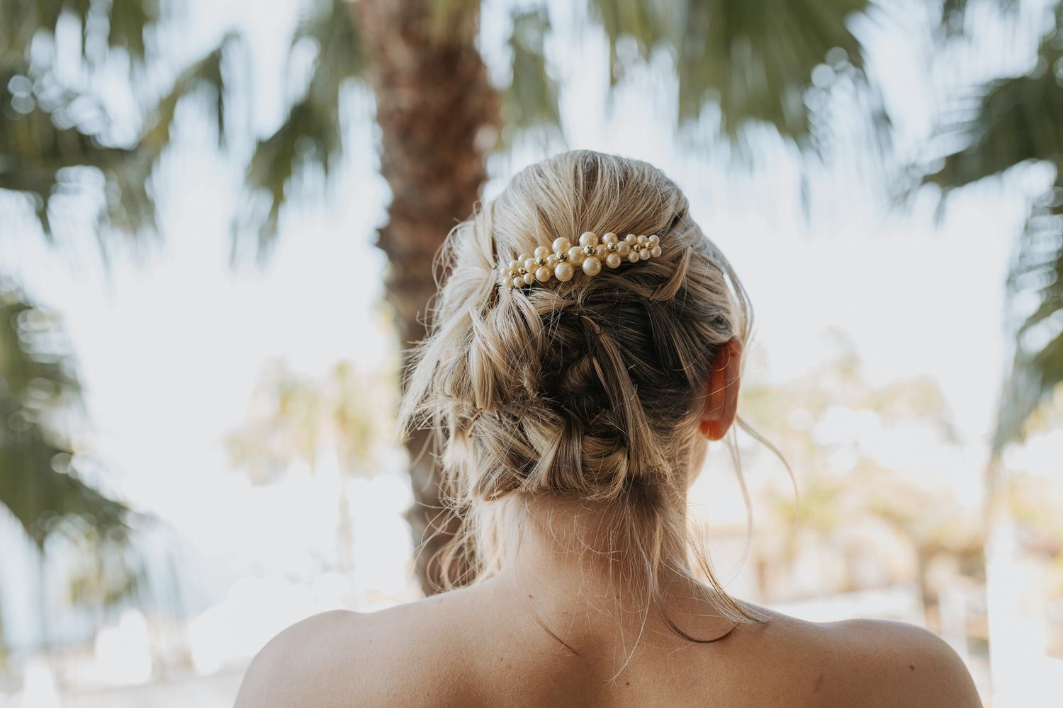 Back view of an elegant bridal hairstyle with delicate gold hair accessory against a palm tree background.