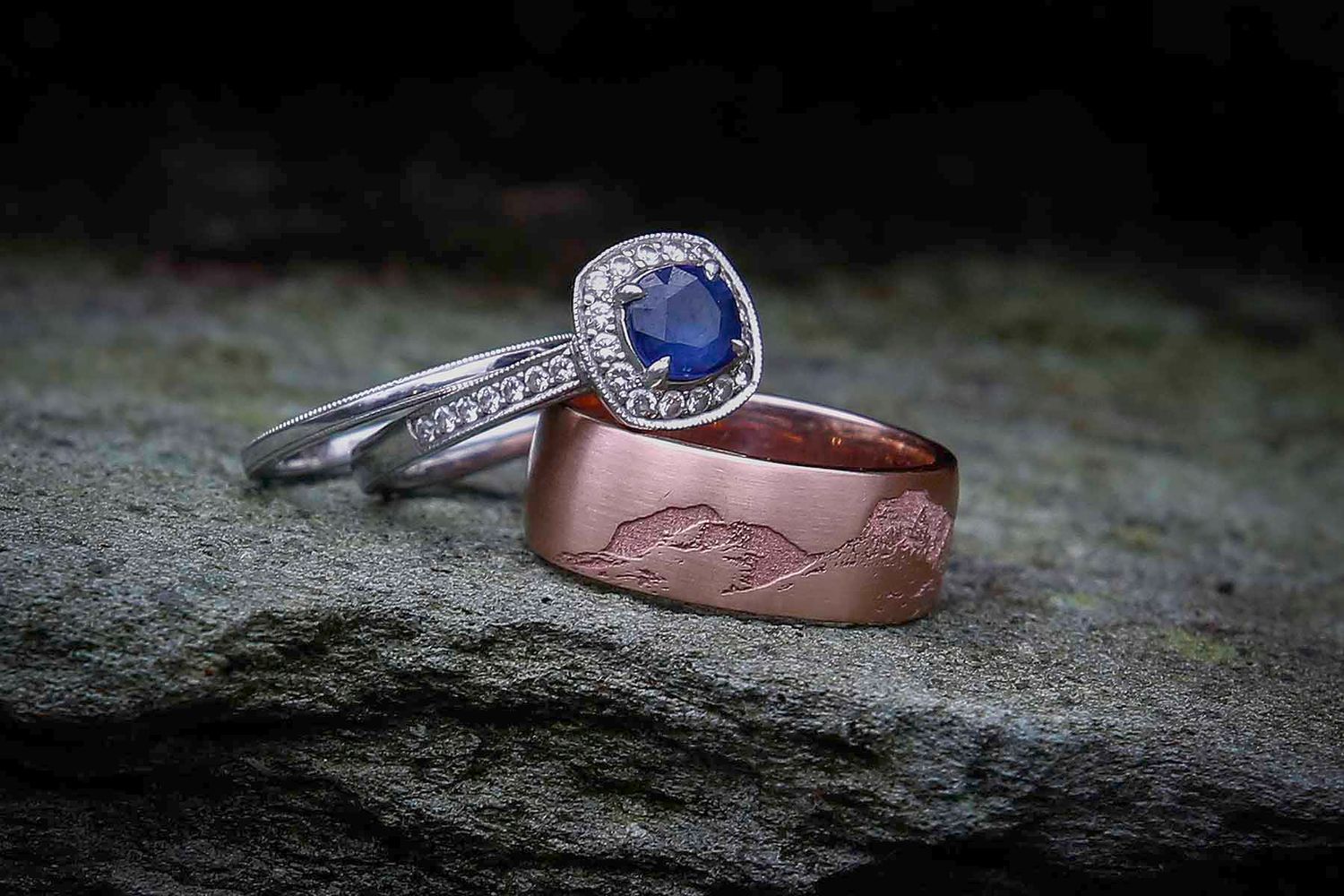 A sapphire engagement ring and rose gold wedding band with engraved design of the langdales in the lake district ,resting on blue slate.