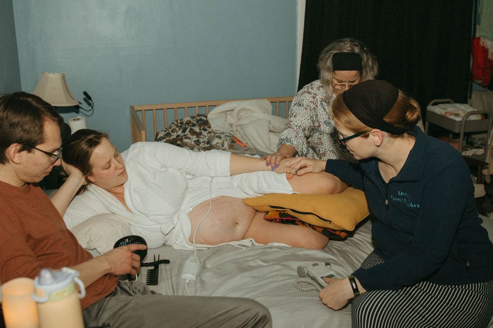 Midwives and support person assist pregnant woman during active home birth labor on bed.