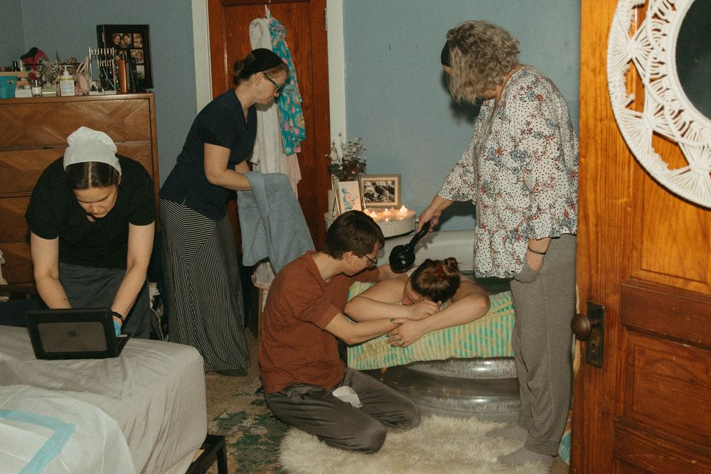 A birth team assists a laboring woman in a home birth setting with candles nearby.