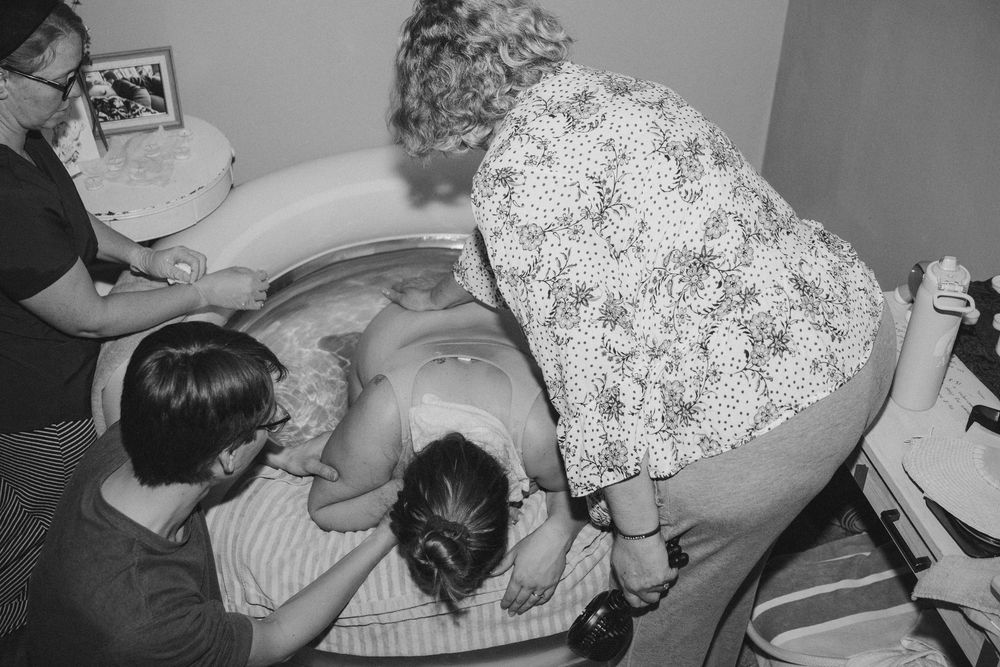 Black and white image of a woman laboring over a birth tub supported by a birth team.