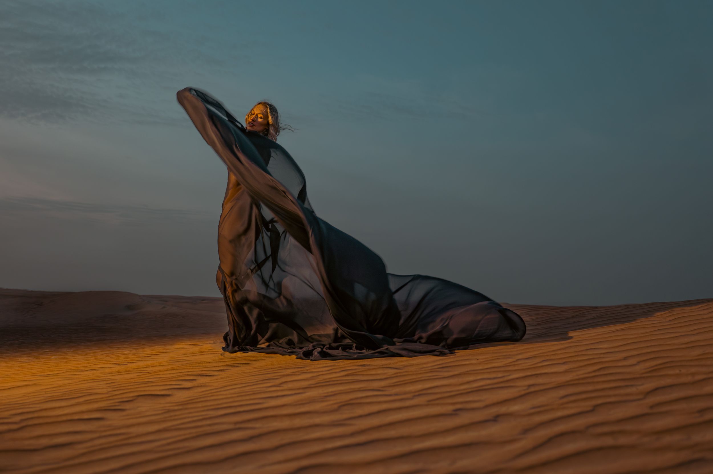 Woman draped in flowing dark fabric kneeling on golden sand dunes at dusk in a serene desert landscape.