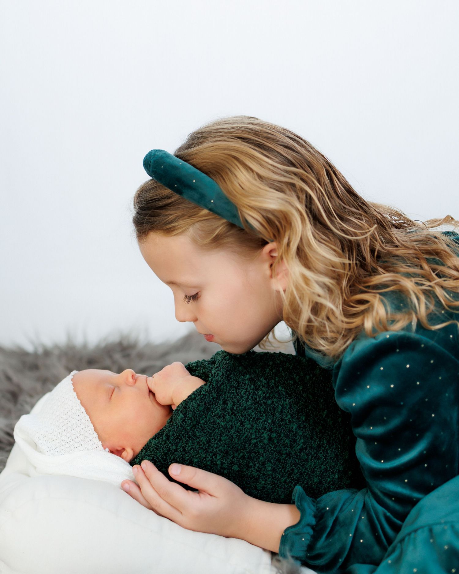 A woman in a teal sweater leans down to kiss a newborn baby wrapped in white blankets against a snowy backdrop.