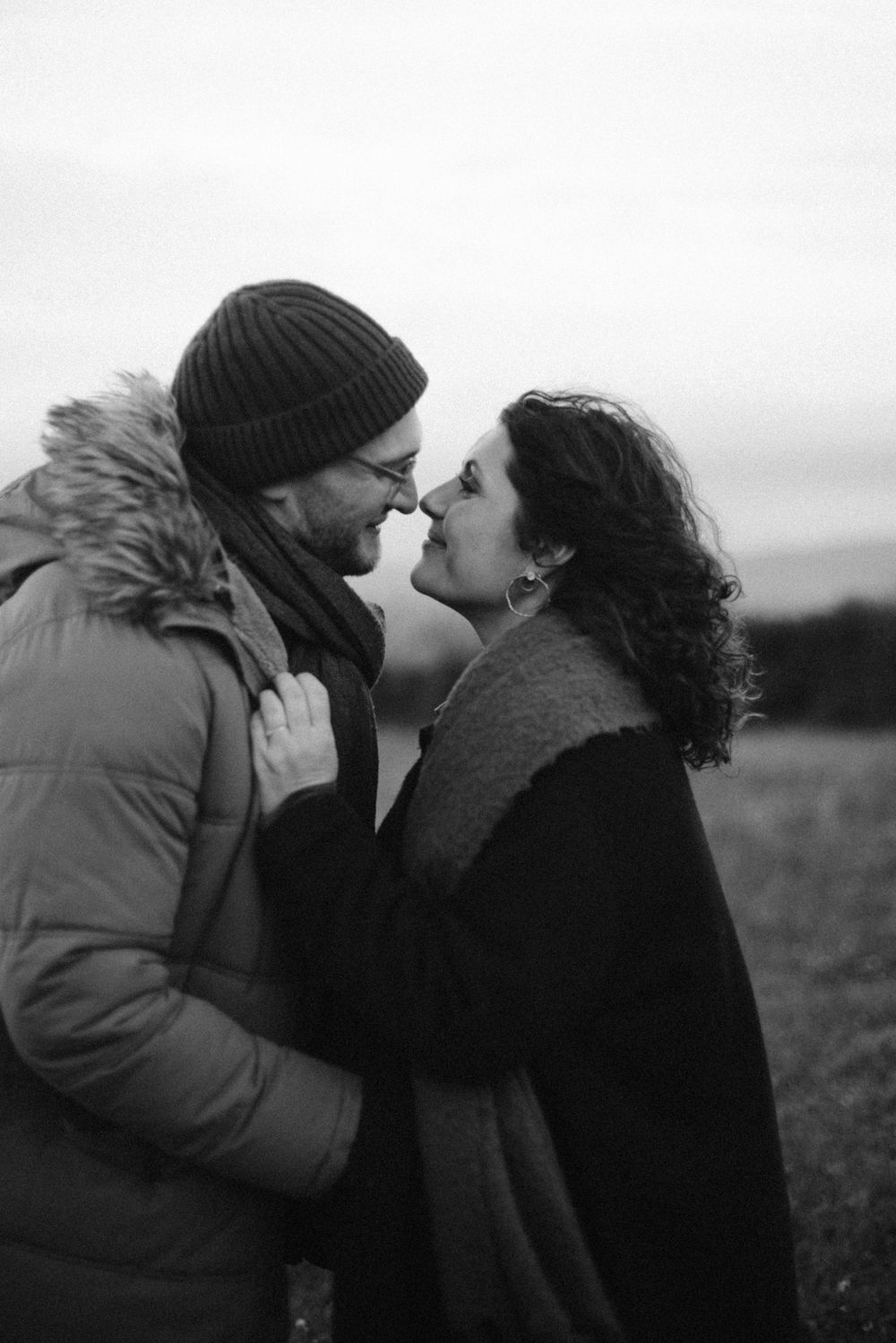 A romantic black and white photo series shows a couple sharing tender kisses in nature while wearing winter clothing.