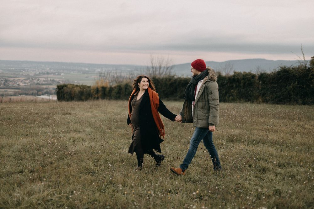 Two people walking through a grassy field on an overcast day with mountains visible in the distance.