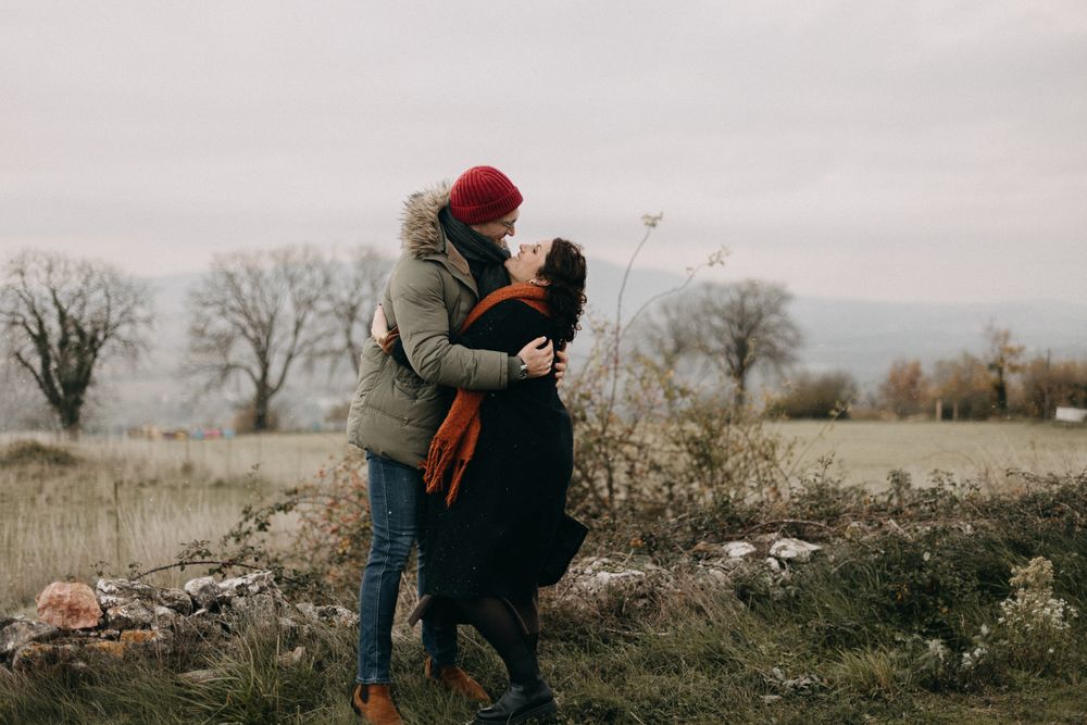 A couple embraces in a winter field with bare trees and dried grass under an overcast sky.