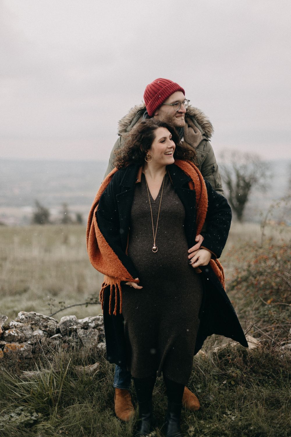 A person wearing a red beanie and brown scarf poses outdoors in autumn against a moody overcast landscape.