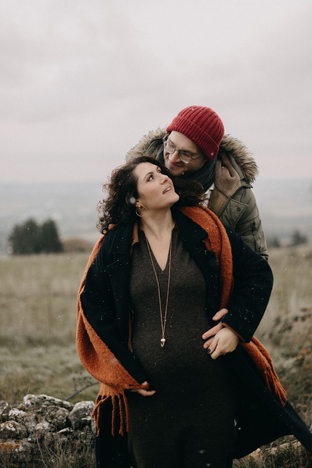 A couple in winter attire embraces on a stone wall in a moody outdoor setting with overcast skies.