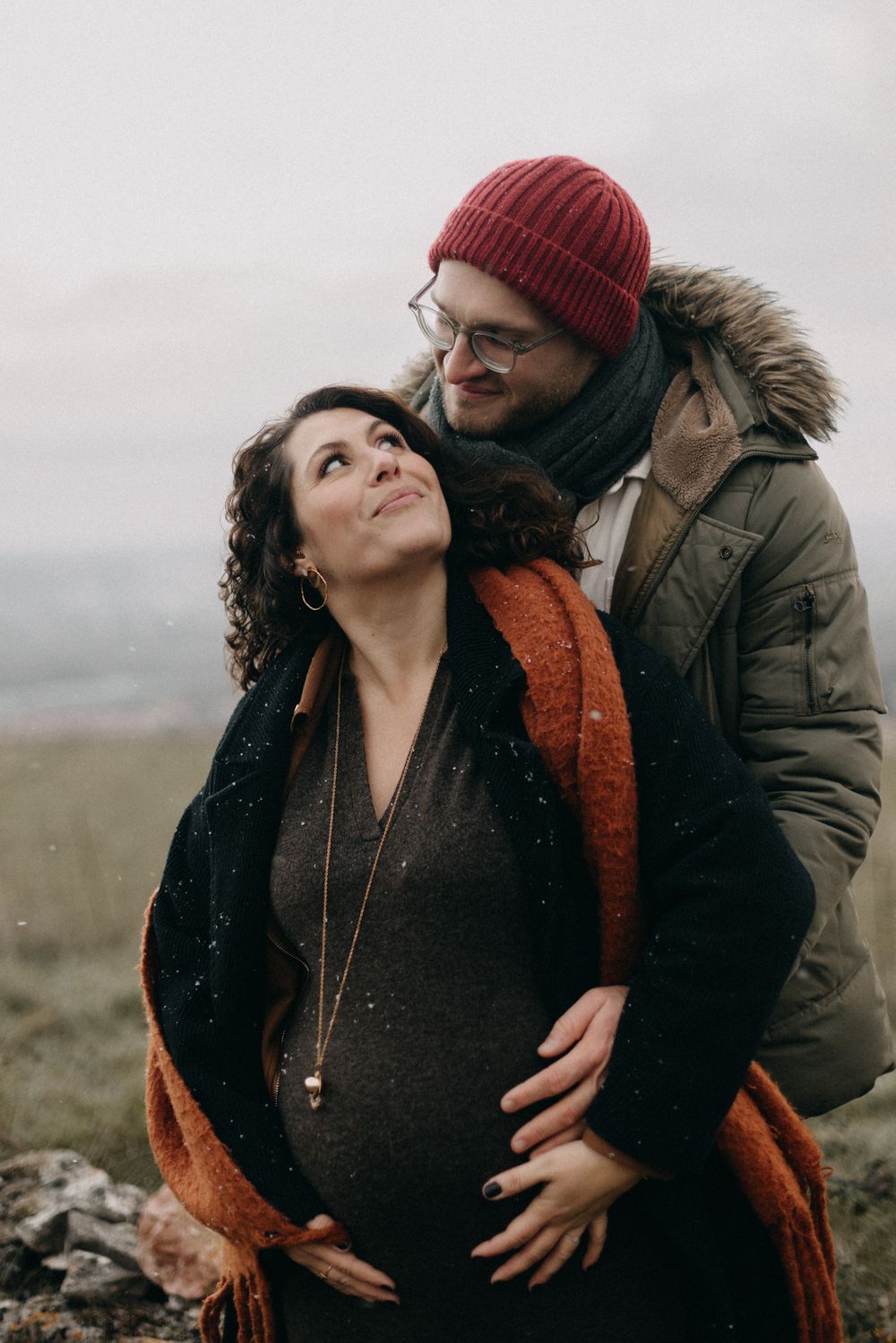 Expectant couple sharing an intimate moment outdoors with the person in a red beanie embracing from behind.