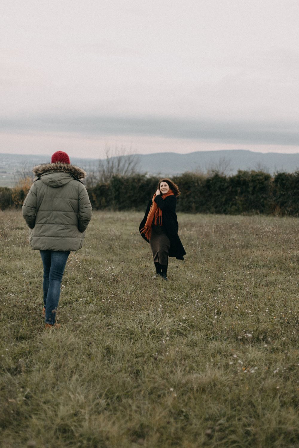 A person in a puffy jacket walks across a brown grassy field on an overcast winter day with mountains in the distance.