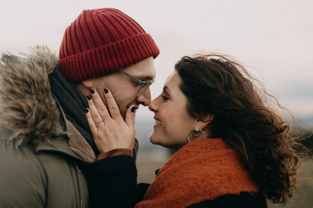 A couple in winter clothing share an intimate moment outdoors, with one wearing a red beanie and the other in an orange sweater.