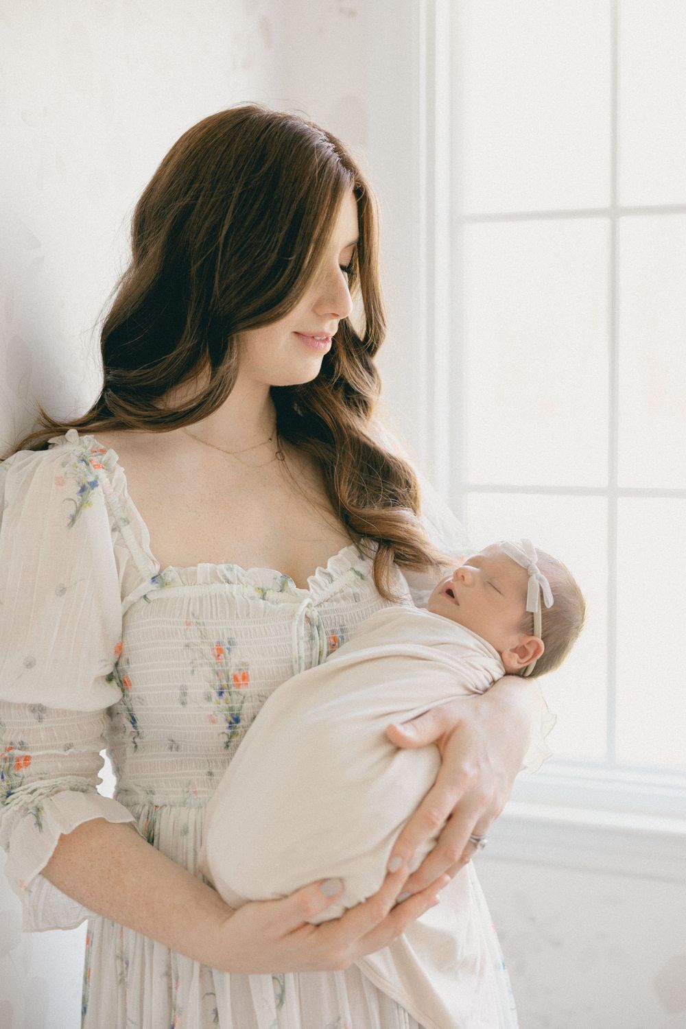 Parent in floral dress holding newborn by bright window in white room, baby nursery in-home session.