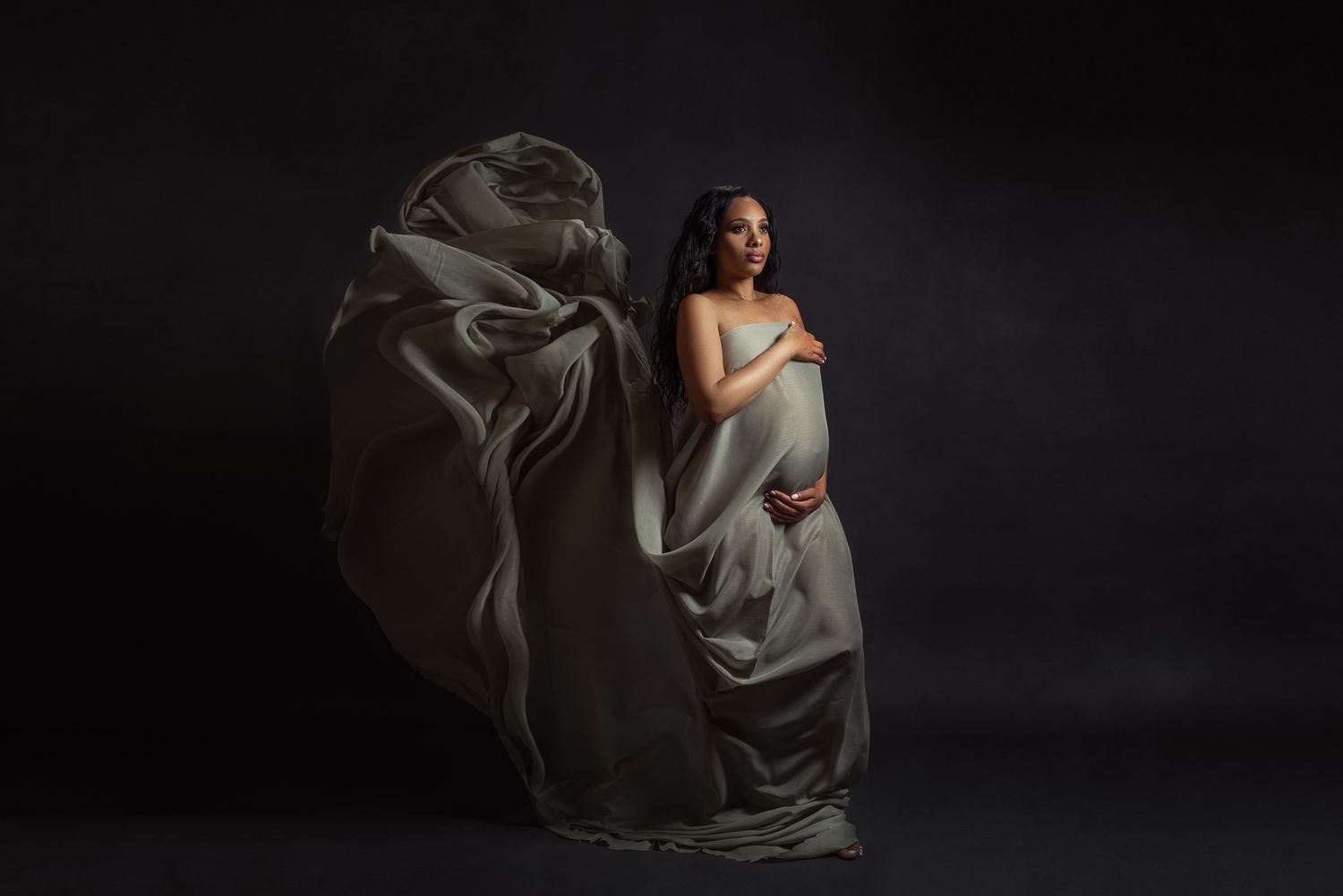 A woman in an elegant blue gown with sheer sleeves and a high slit poses against a dark background in a studio setting.