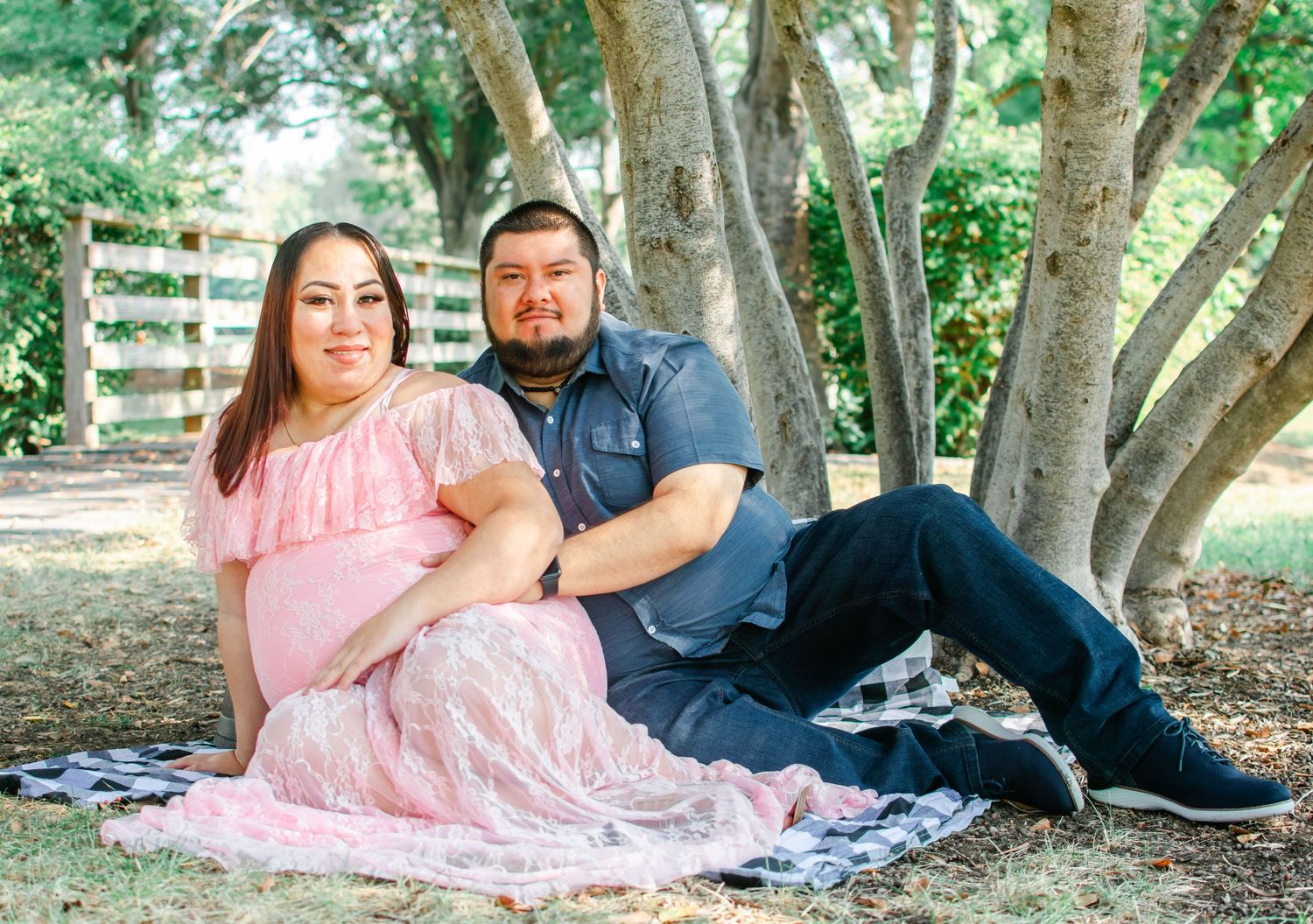 A couple sits together on a blanket under shady trees for a romantic outdoor maternity photoshoot.