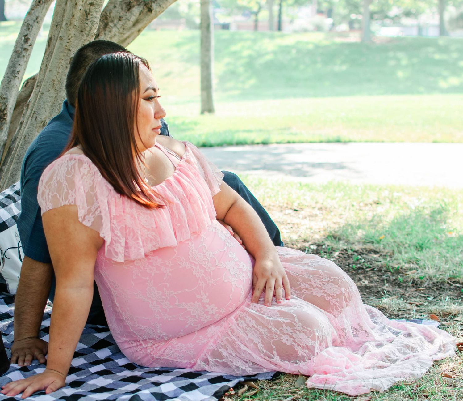 A woman in a pink lace maternity gown poses gracefully on the grass during an outdoor photo session.