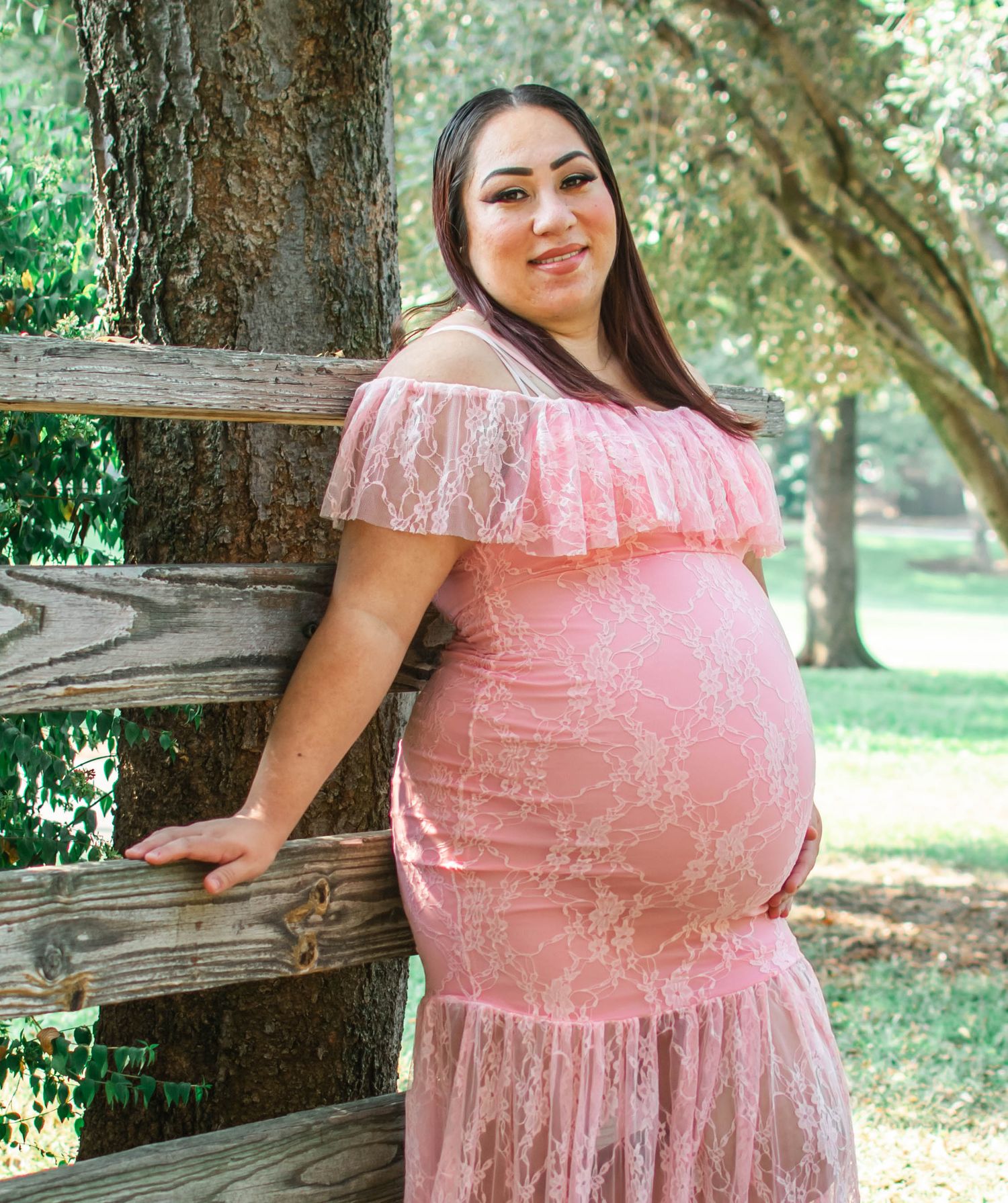 A person in a pink lace dress poses outdoors against a wooden fence in a natural setting.