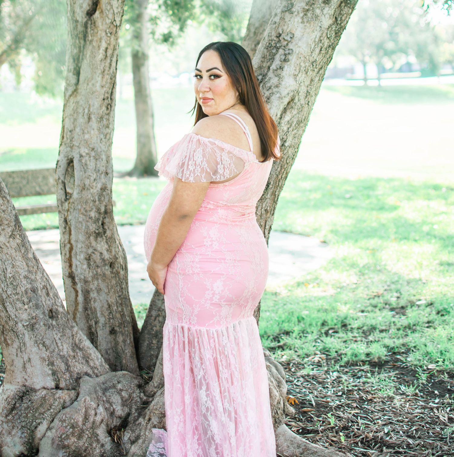 A person in a pink lace dress poses gracefully against a large tree trunk in a sunlit outdoor setting.