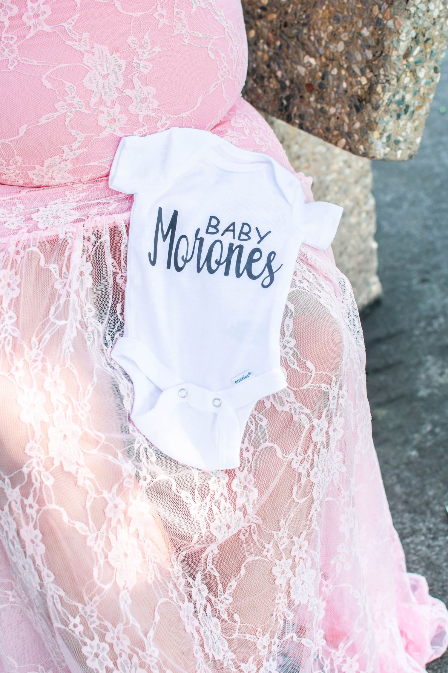 A white baby onesie with 'Baby Morones' text rests on pink lace fabric.