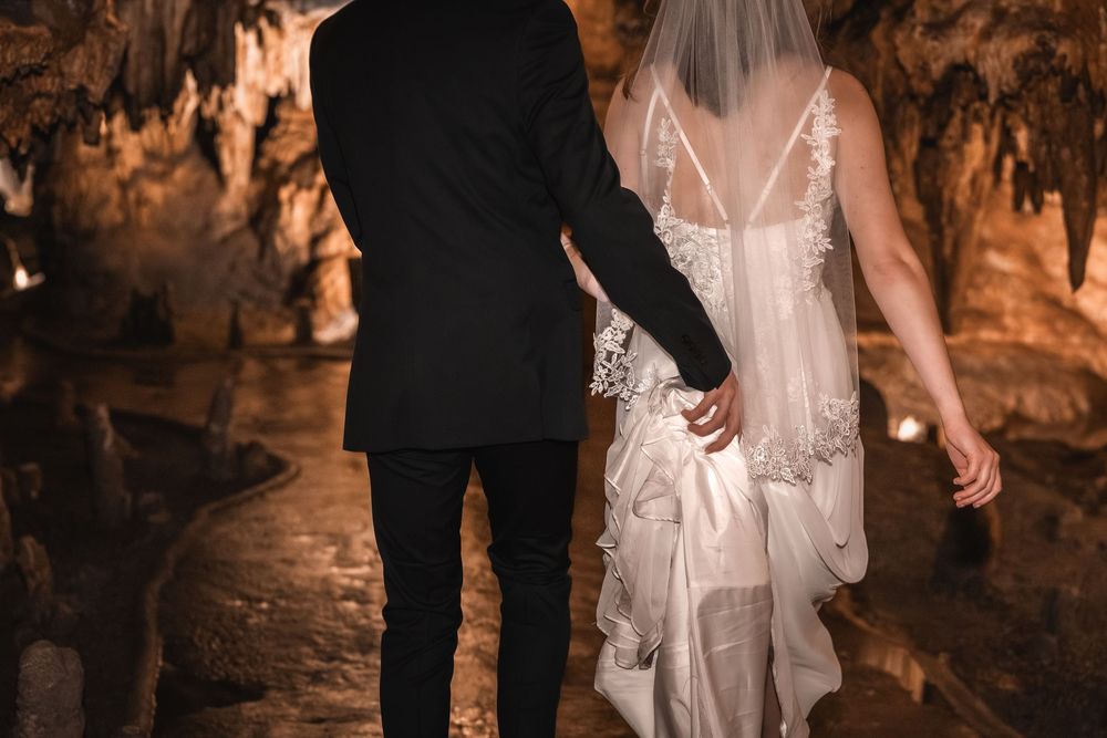Bride and groom hold hands while walking through dimly lit cave formations during wedding photo session.