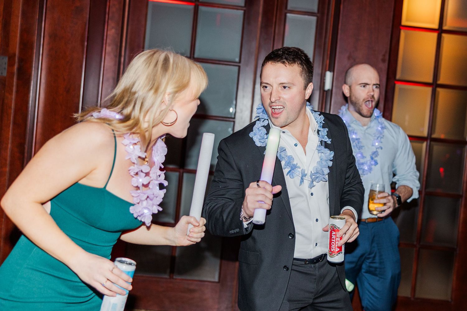 People dancing and celebrating at a wedding reception in formal attire with drinks in hand.