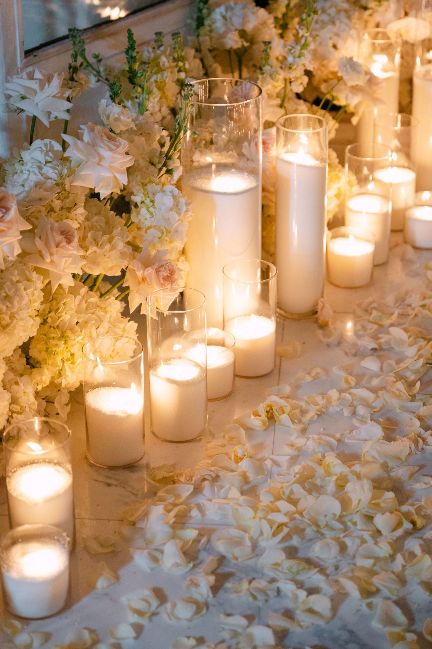 White pillar candles glowing warmly among scattered flower petals and greenery create an elegant wedding reception display.