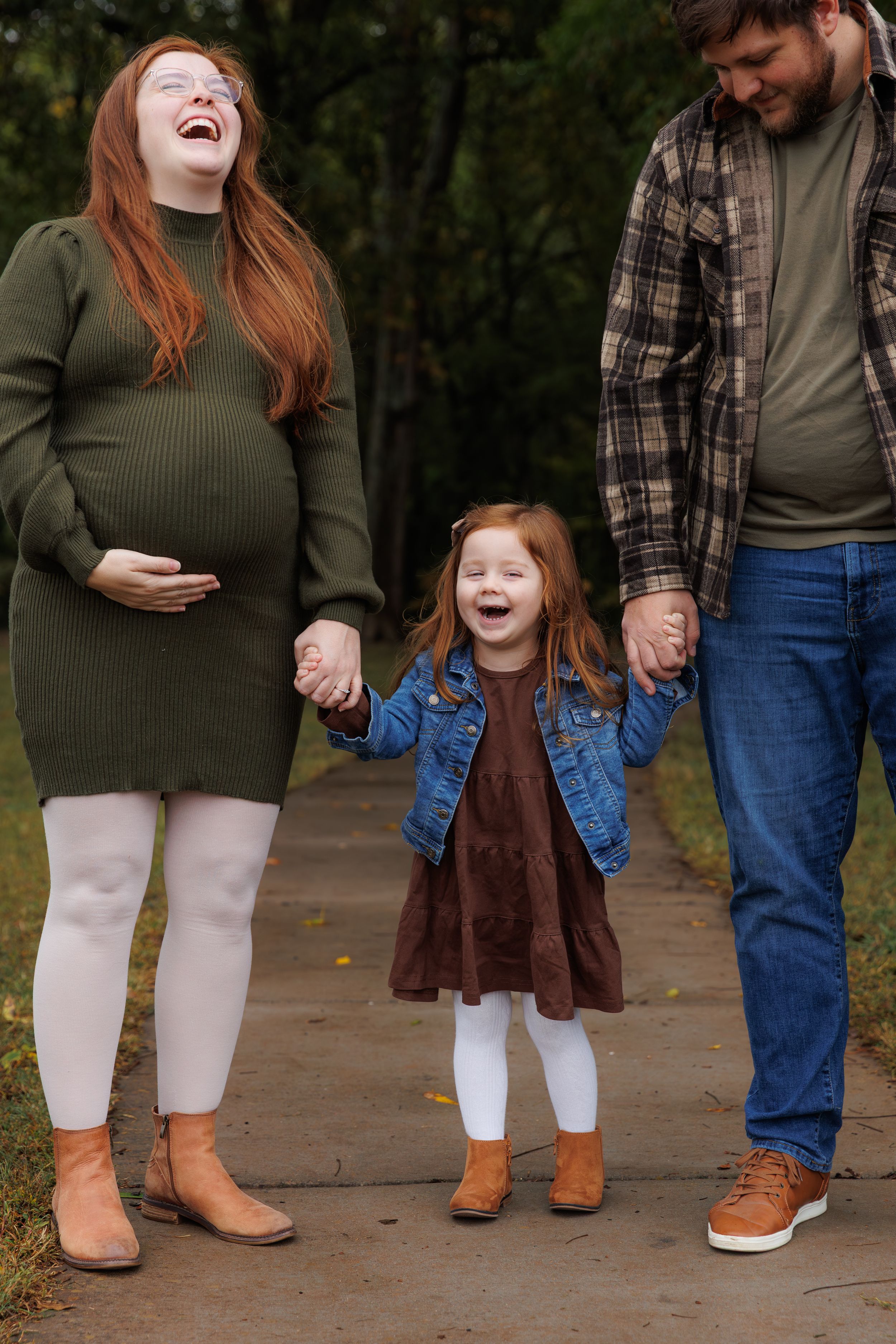 A family laughs together while holding hands on an autumn path, with a pregnant person in green and a child in denim.