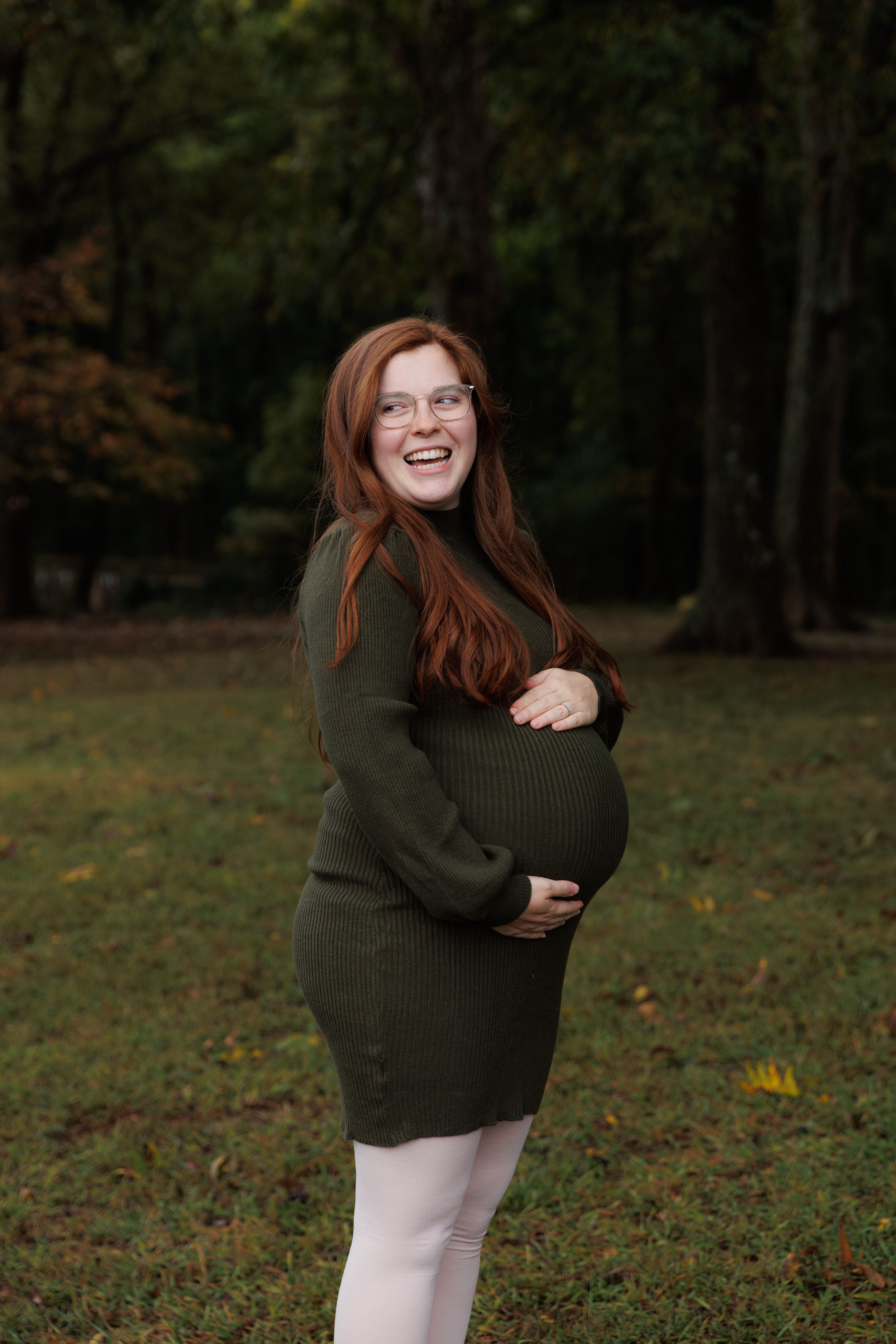 A person in a green sweater and white pants poses playfully in an autumn forest setting.