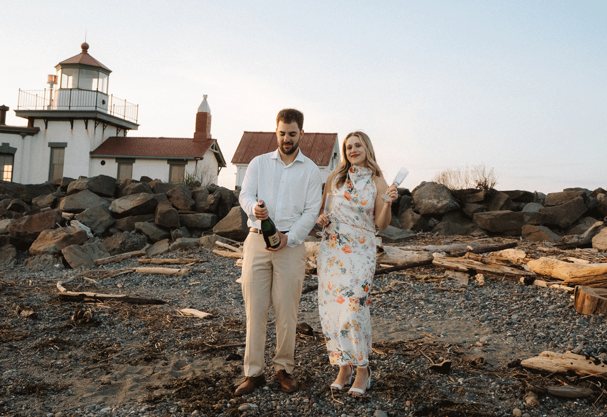 A couple in white outfits walking along a rocky beach with a lighthouse in the background at sunset.