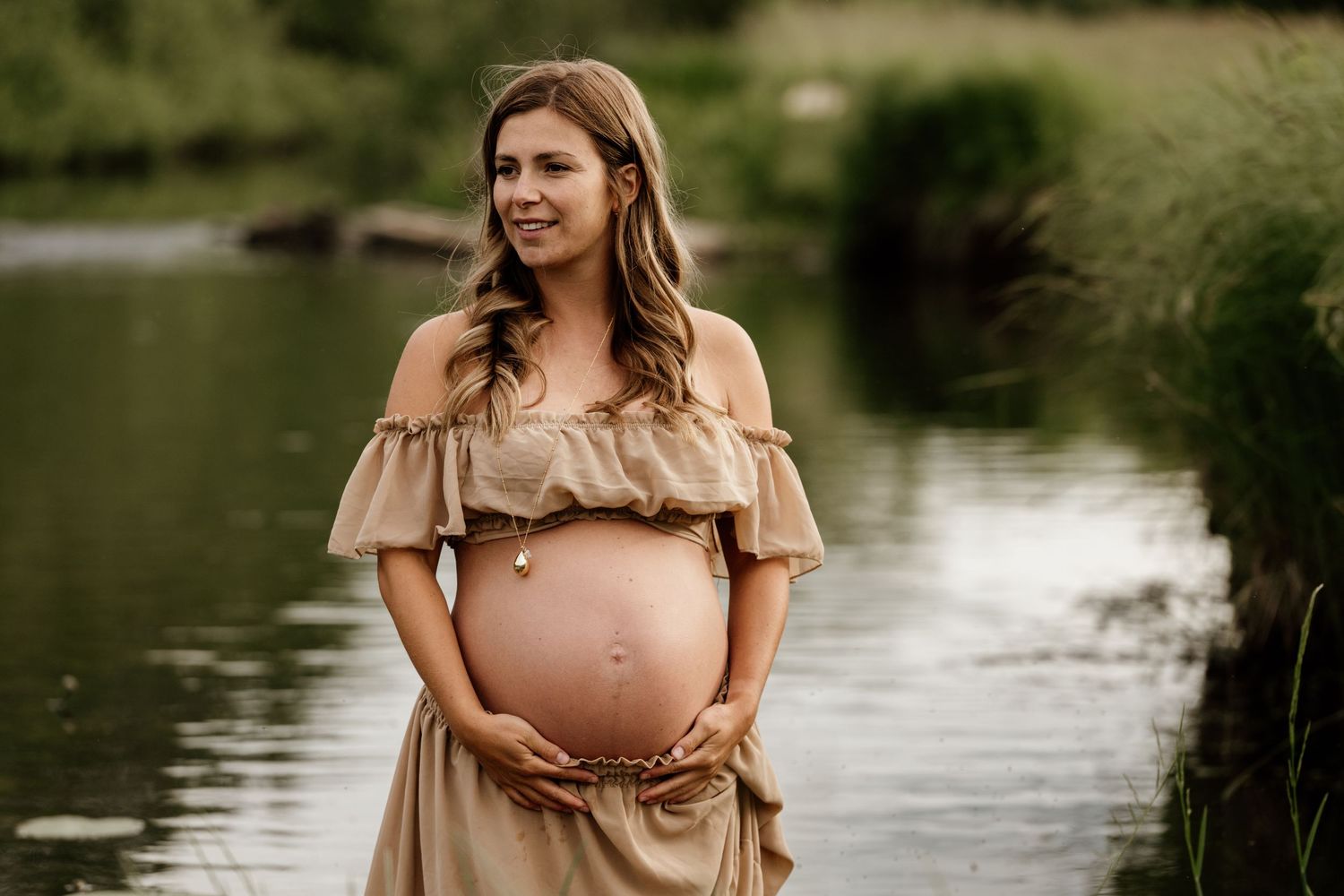 Pregnant woman in a beige off-shoulder gown standing by a reflective pond at golden hour.
