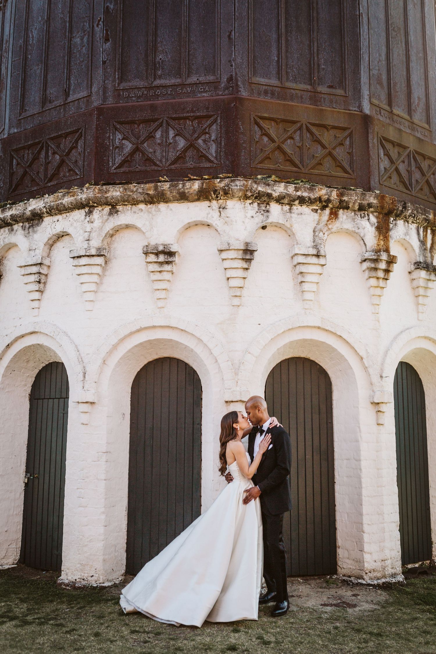 A couple in wedding attire embrace against a historic white stone building with arched doorways and ornate architectural details.