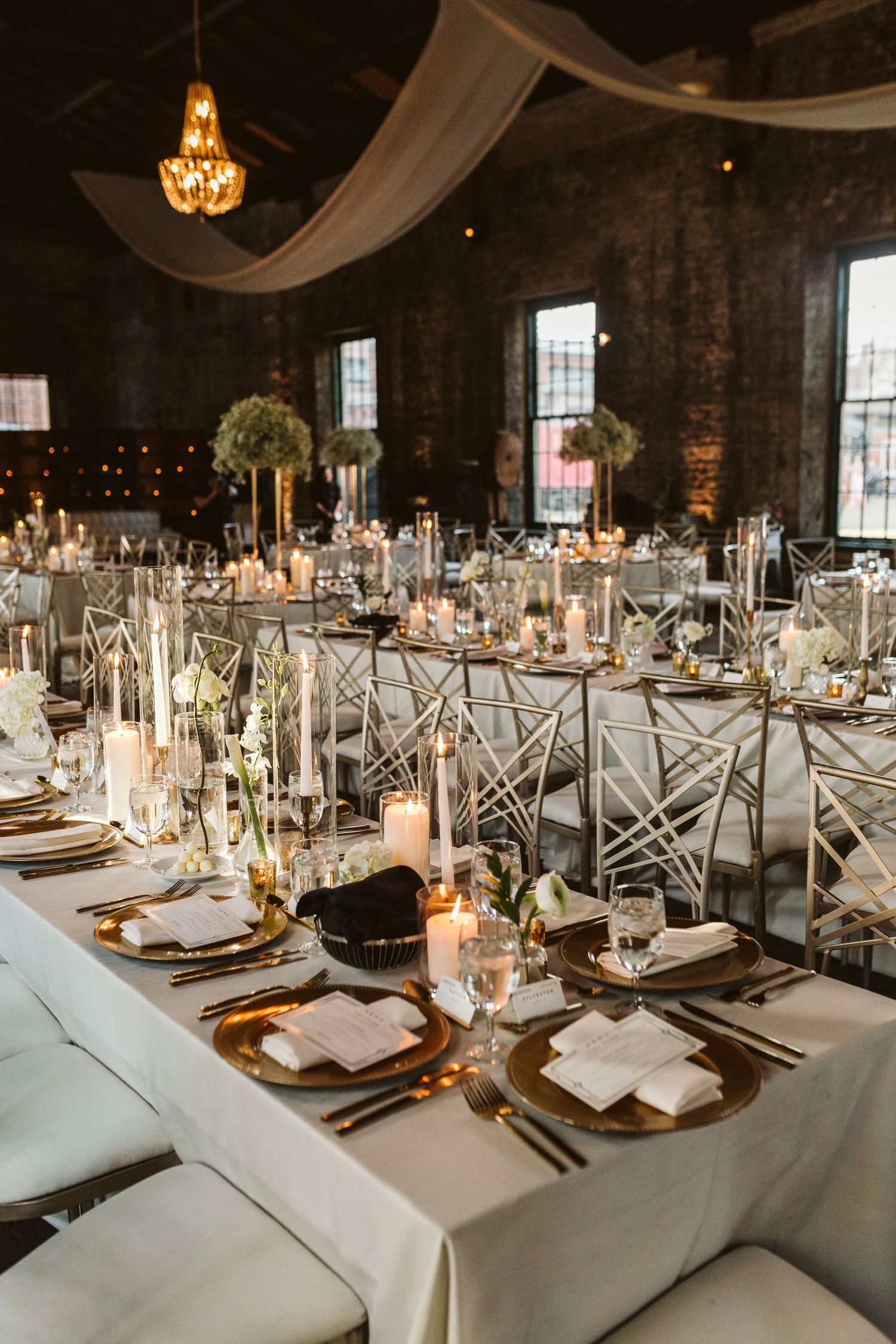 Elegant wedding reception tables set with white linens, gold chargers and crystal glassware in an industrial-chic venue.