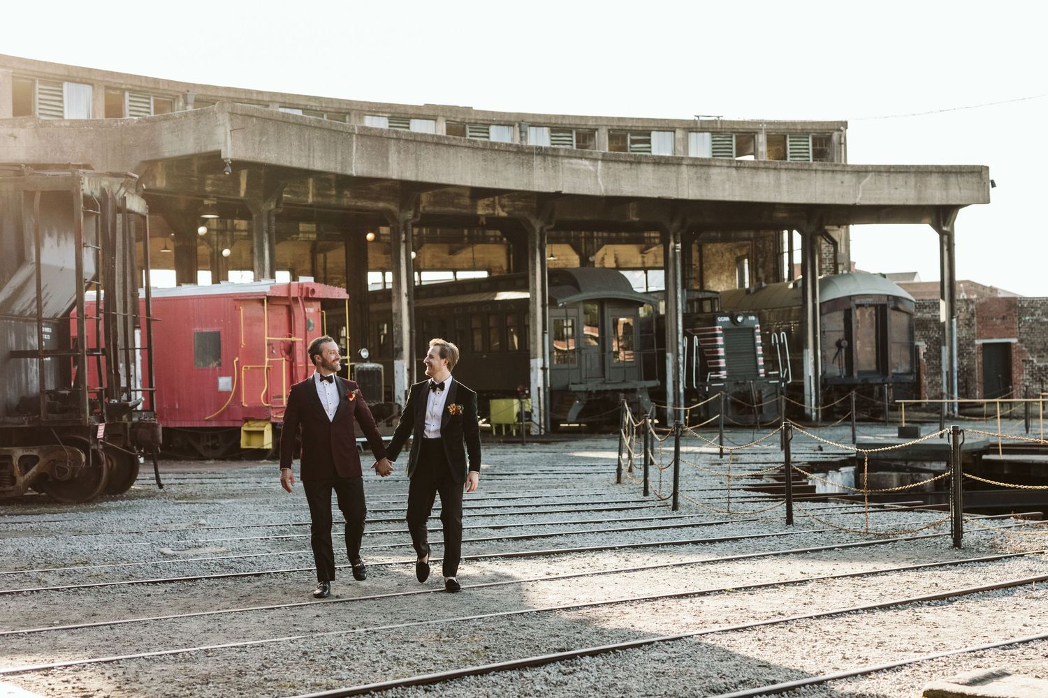 Two people walk along a historic train depot platform with vintage railcars and industrial architecture in the background.