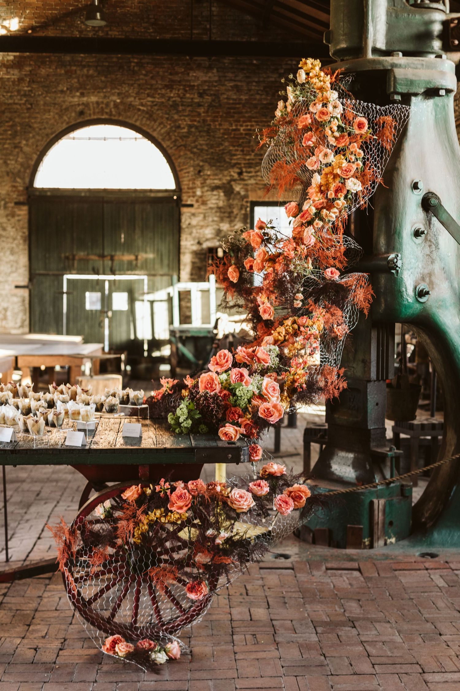 A rustic brick venue interior decorated with peach and coral floral arrangements on wrought iron stands.
