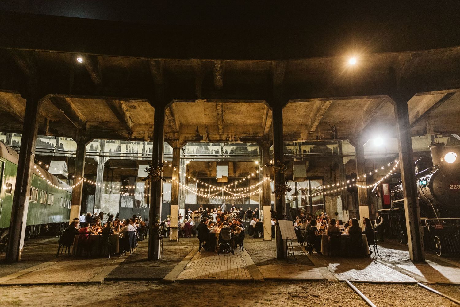 String lights illuminate a rustic outdoor venue with wooden beams and tables set up for an evening event.