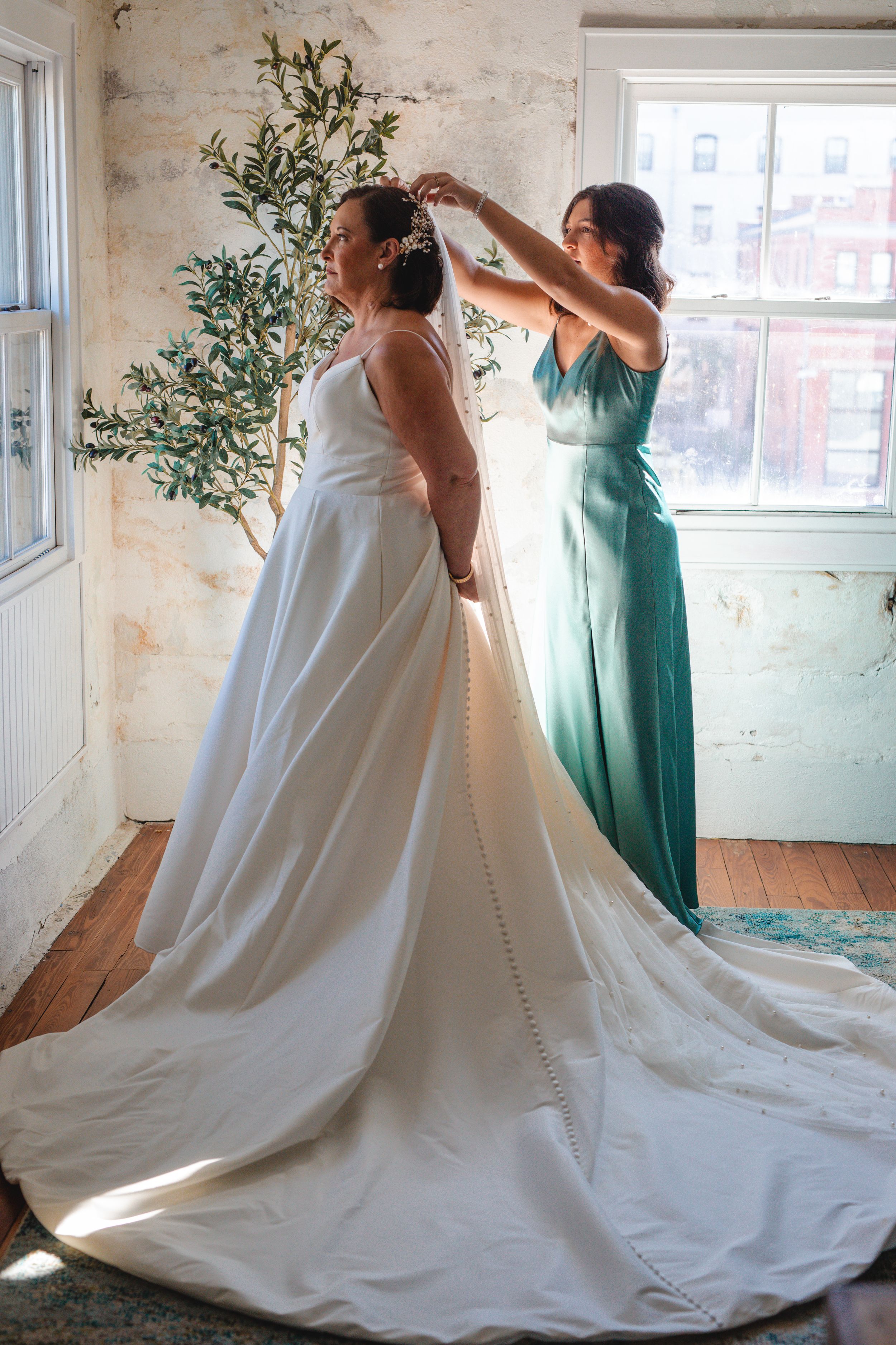 Bridesmaids in mint green dresses help adjust a flowing white wedding gown with a dramatic long train in a sunlit room.
