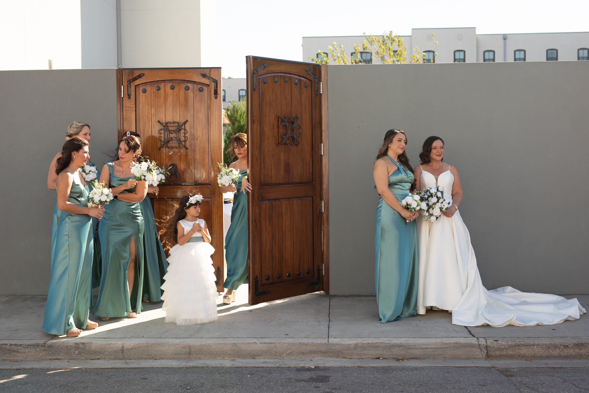 Wedding party poses in coordinated teal and white attire in front of a rustic wooden door and stone wall.