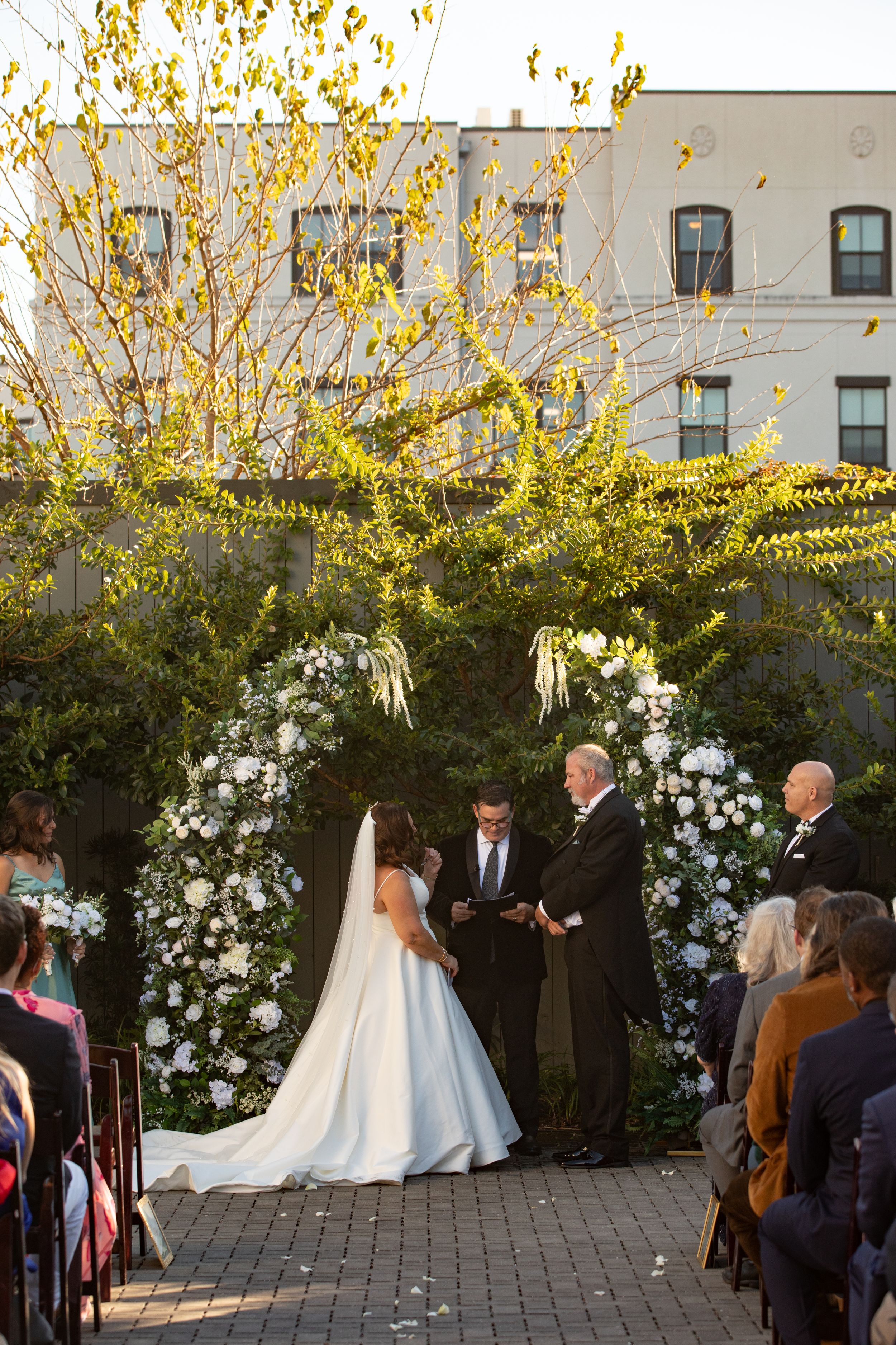 Outdoor wedding ceremony under a floral arch with white roses and golden autumn foliage at sunset.
