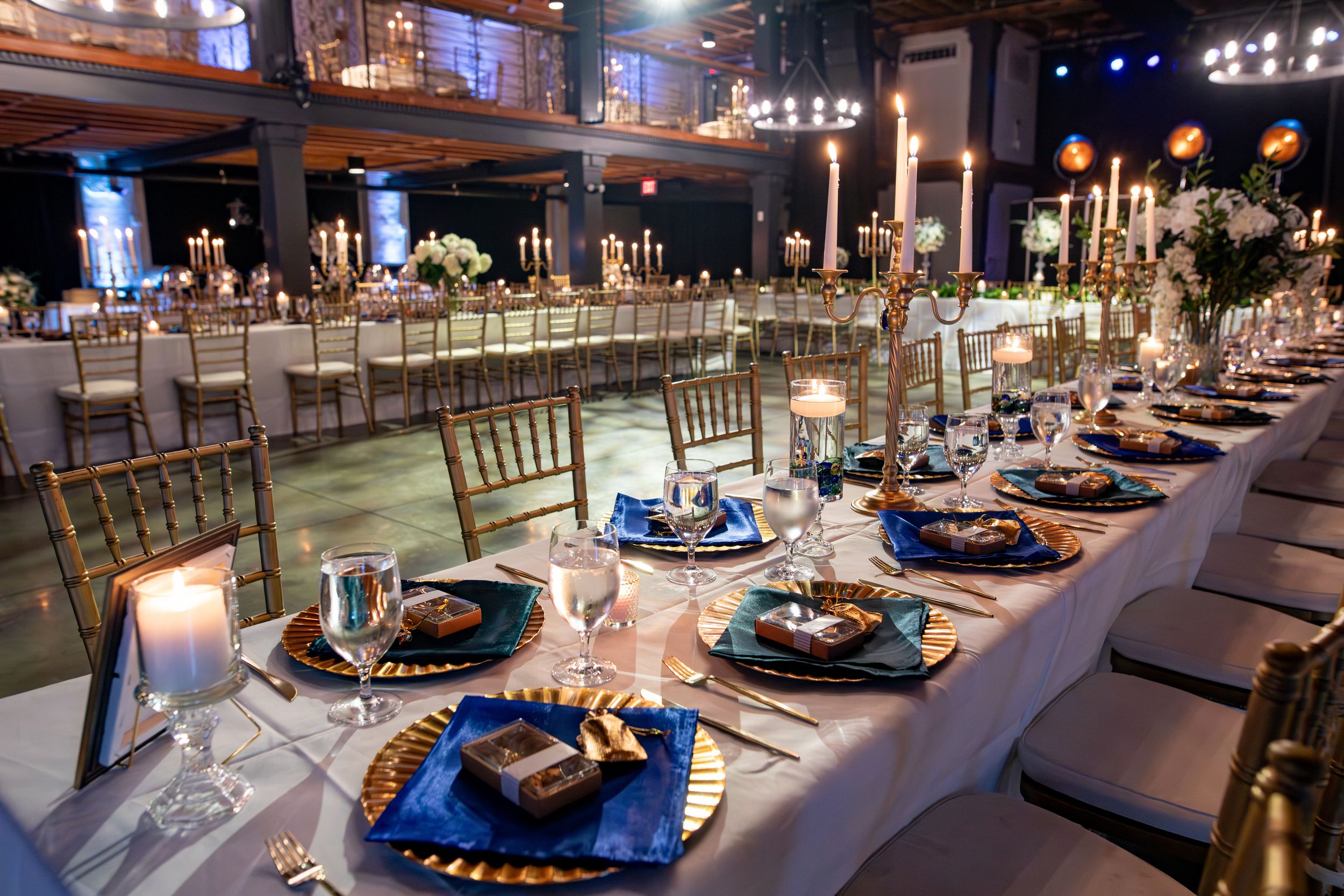 Elegant wedding reception venue with blue uplighting and formal table settings featuring navy and gold decor elements.