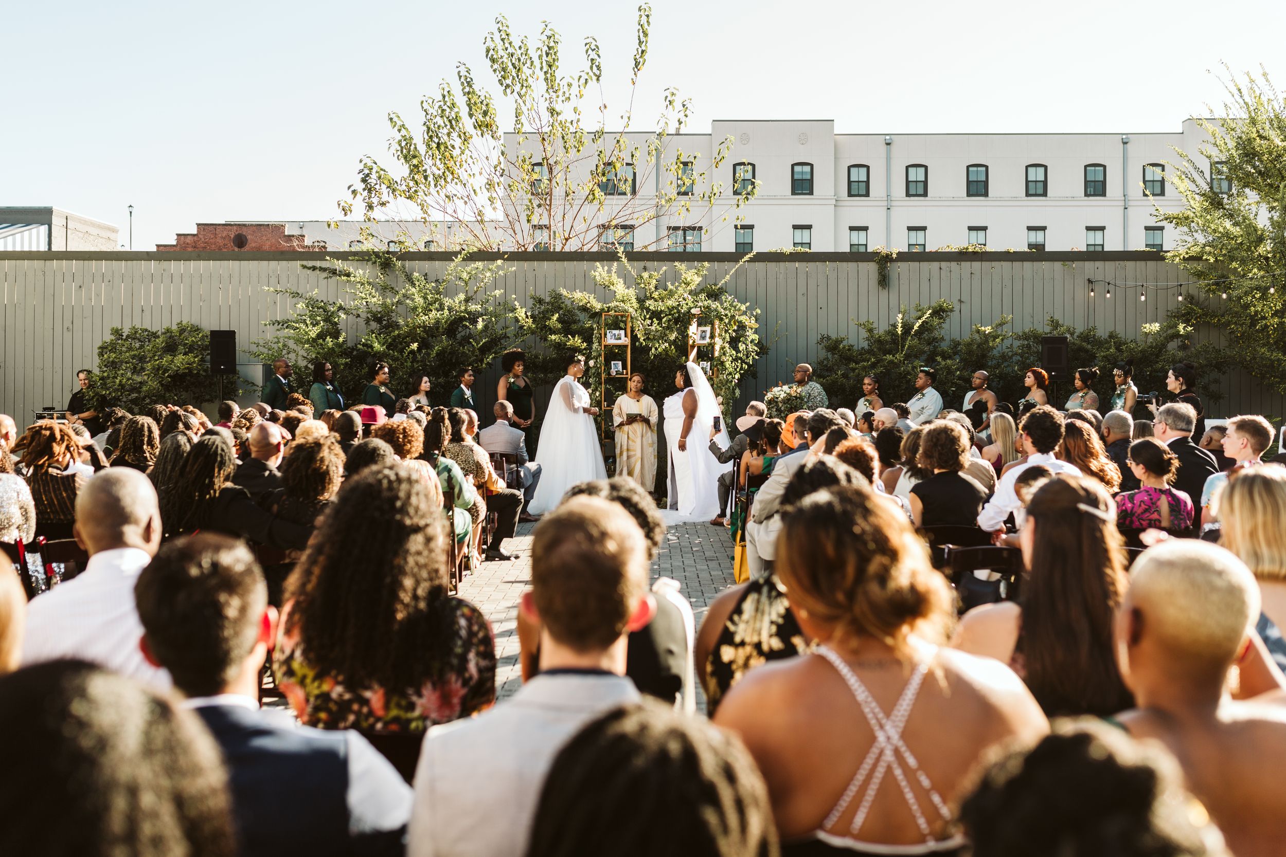 A large crowd of wedding guests seated outdoors watching a ceremony in progress on a sunny day.