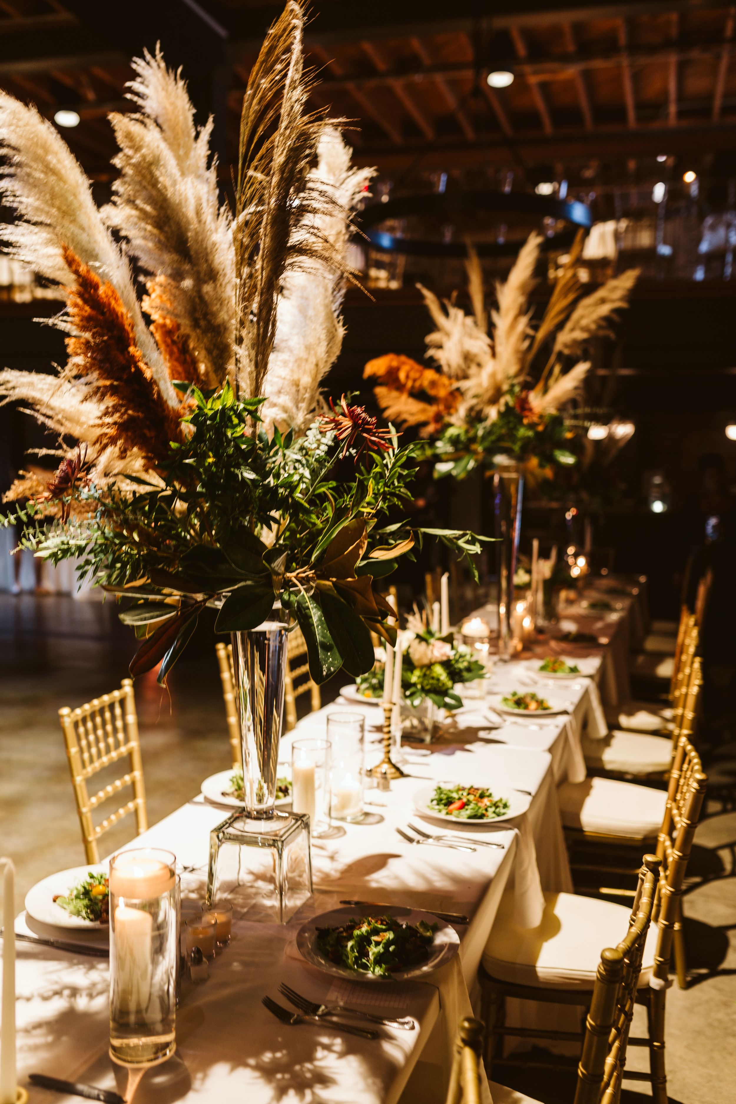 Elegant wedding reception table setting with tall pampas grass centerpieces and gold chiavari chairs in dim lighting.
